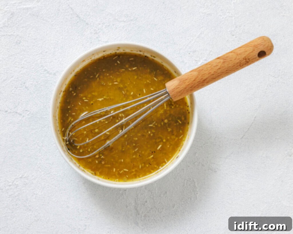 A bowl of yellow-green vinaigrette dressing with visible herbs, with a metal whisk resting inside.