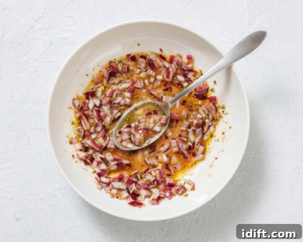 A white bowl with chopped red onions, oil, herbs, and vinegar being mixed with a spoon on a light surface.