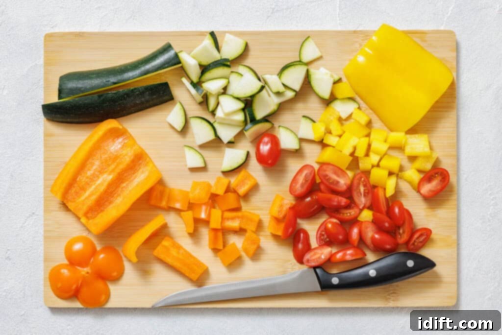 Chopped zucchini, yellow bell pepper, orange bell pepper, and halved cherry tomatoes on a wooden cutting board beside a knife.