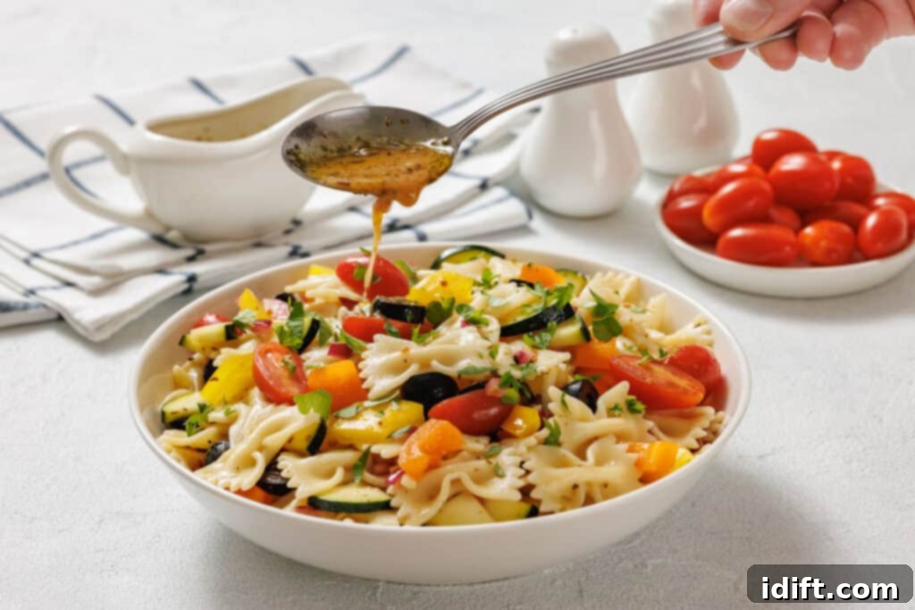 A hand drizzles dressing over a bowl of pasta salad with cherry tomatoes, zucchini, bell peppers, and herbs. A bowl of tomatoes, sauce, and salt and pepper shakers are in the background.