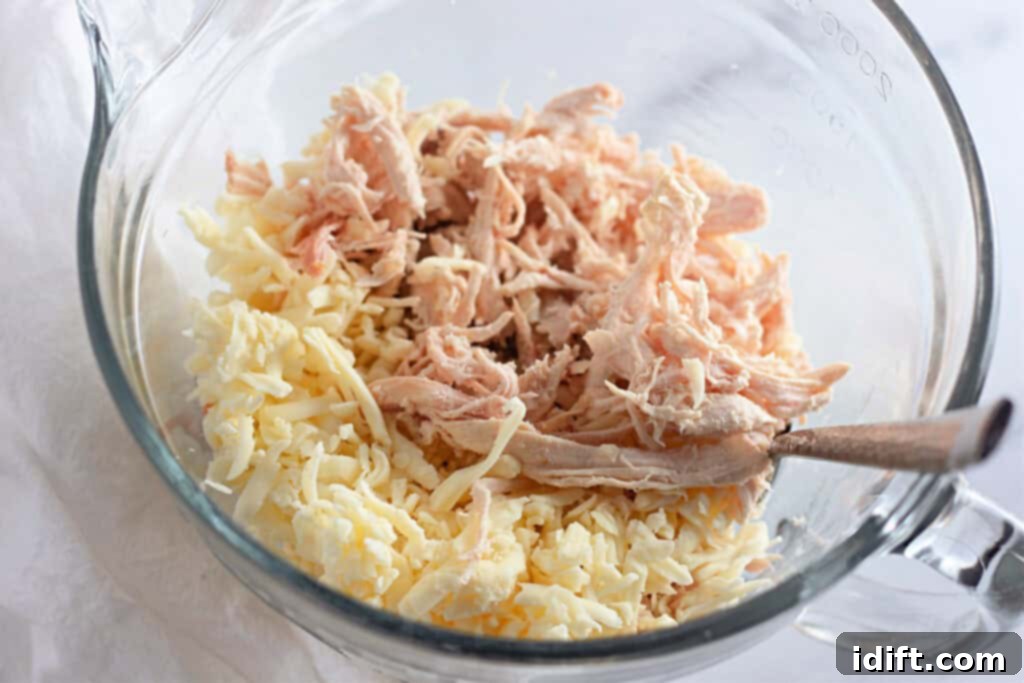 Velvety Chicken Enchiladas 5 Shredded chicken and grated Monterey Jack cheese are mixed in a clear glass bowl, with a serving spoon resting beside it, ready for filling tortillas.