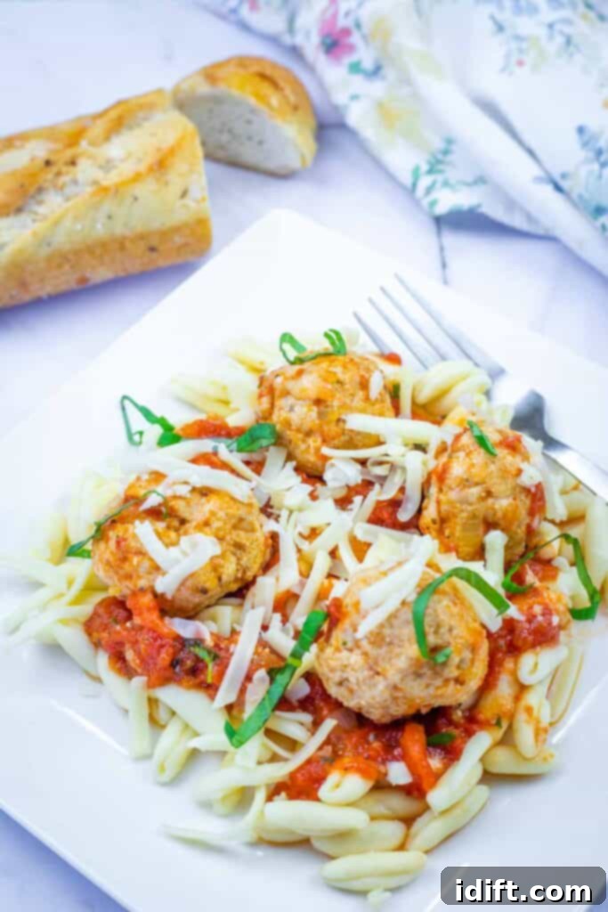 A plate of pasta topped with tomato sauce, meatballs, shredded cheese, and basil, served with a fork and a slice of bread in the background.