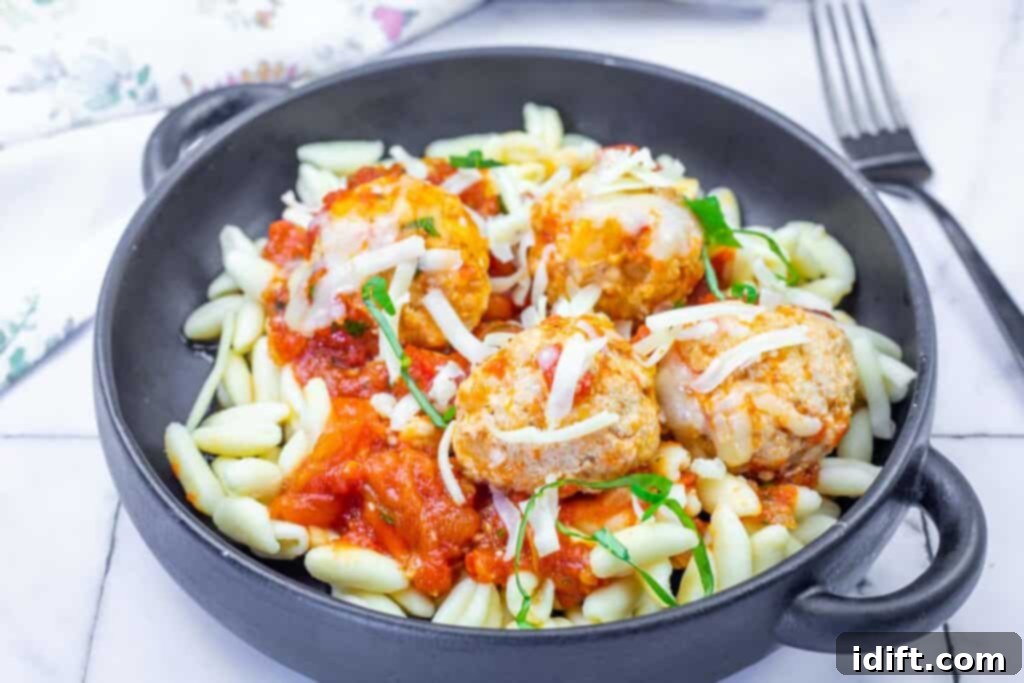 A black bowl filled with pasta, tomato sauce, and four meatballs topped with melted cheese and chopped herbs, with a fork in the background.