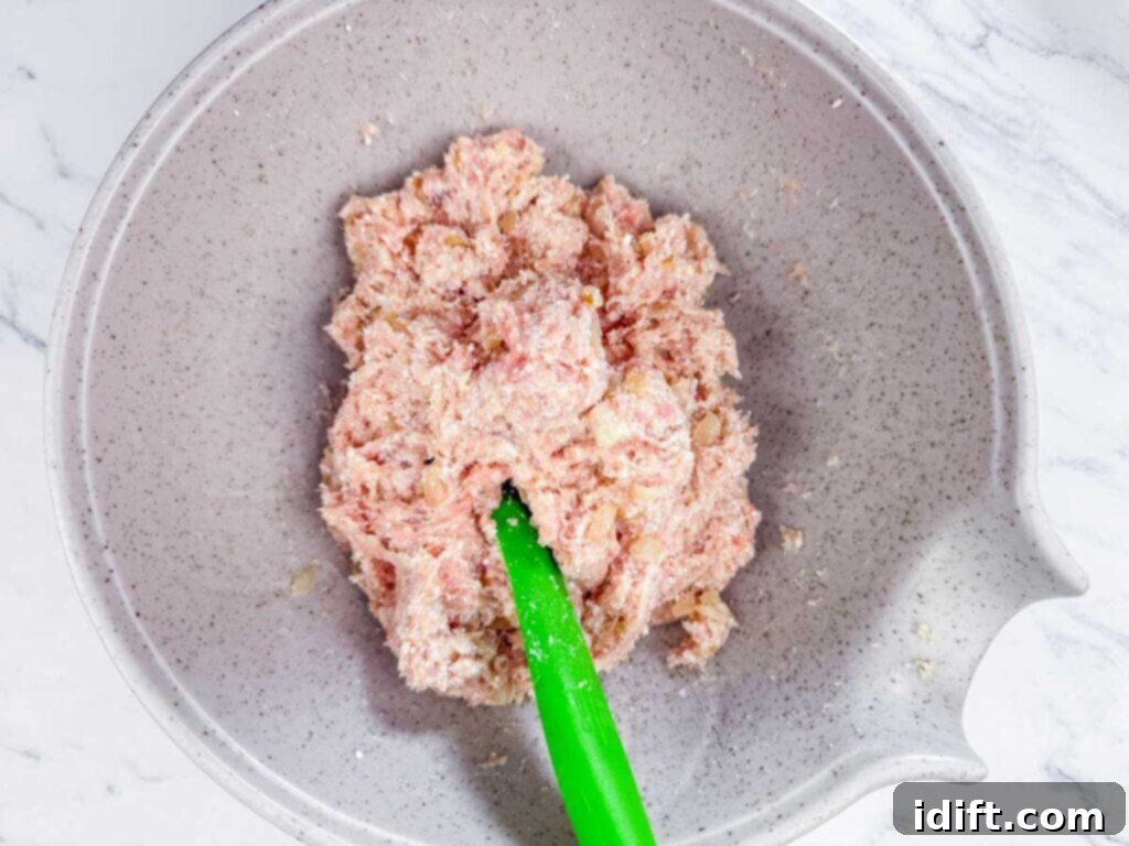 A gray mixing bowl containing raw ground meat mixture being stirred with a green spatula on a white marble surface.