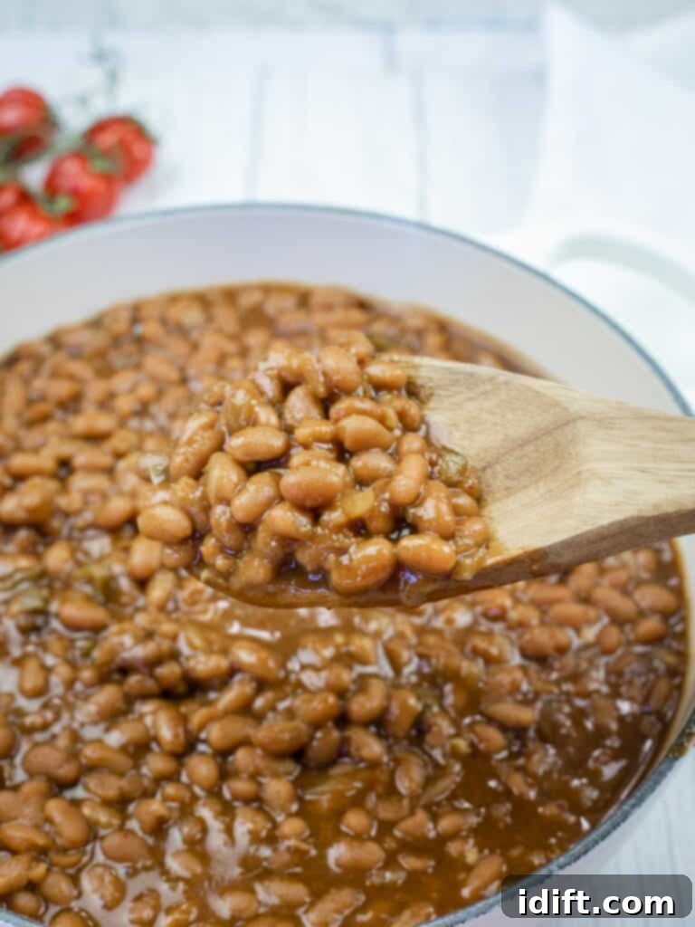 A wooden spoon holding baked beans above a pot filled with baked beans in sauce; tomatoes are visible in the background.
