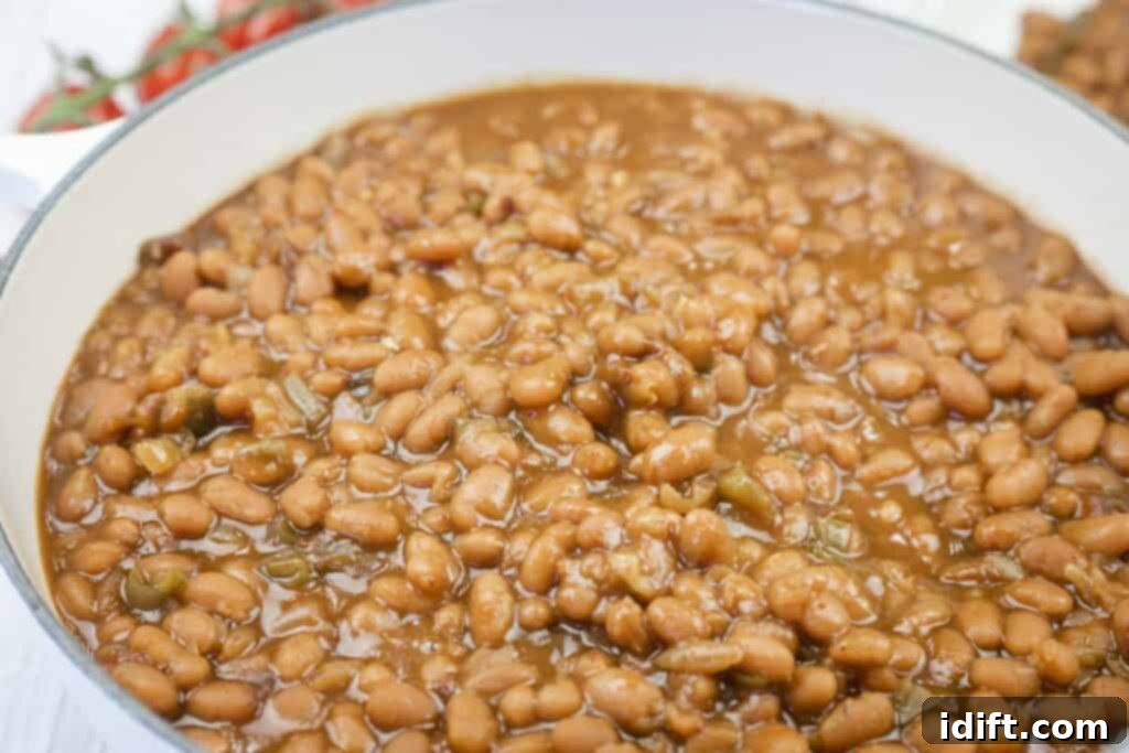 A close-up view of baked beans in a white pot, showing the beans in a thick, brown sauce with visible small pieces of vegetables.