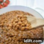 A wooden spoon holding baked beans above a pot filled with baked beans in sauce; tomatoes are visible in the background.