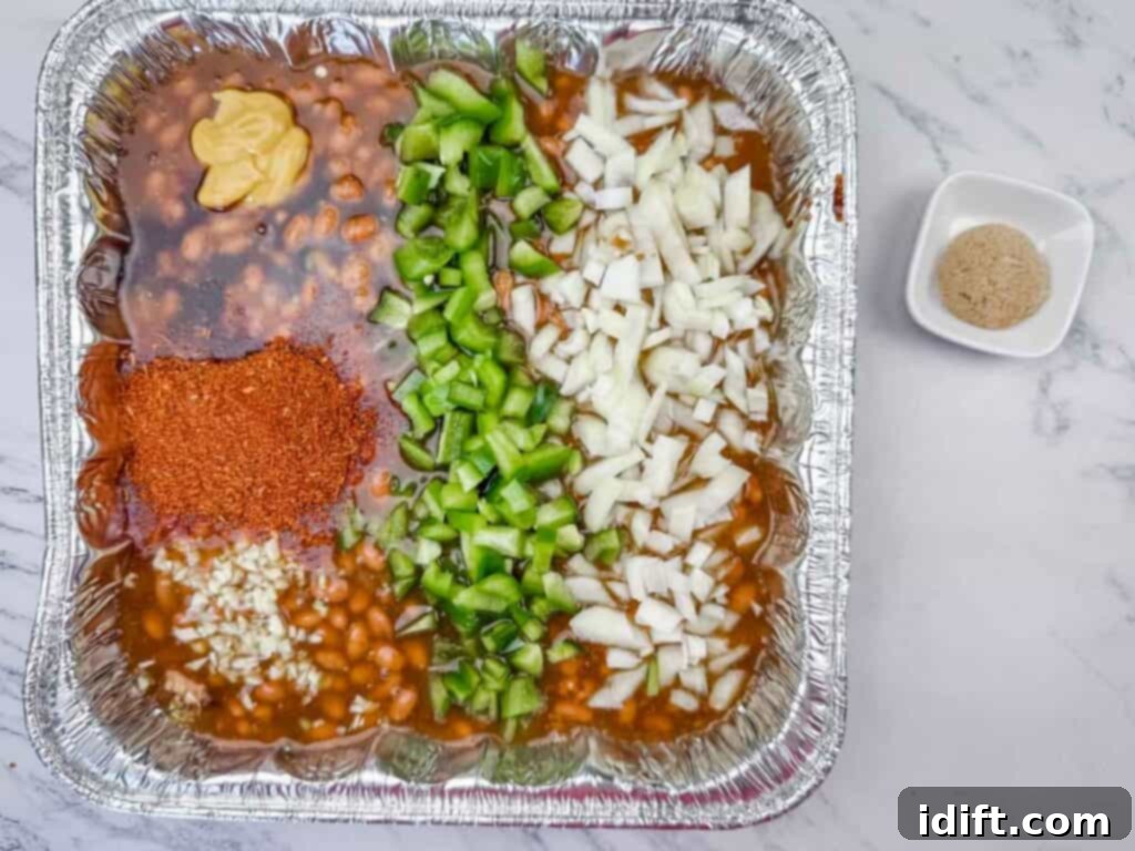 A foil tray with baked beans, chopped green bell peppers, chopped onions, minced garlic, yellow mustard, and chili powder, with a small bowl of brown sugar on the side.