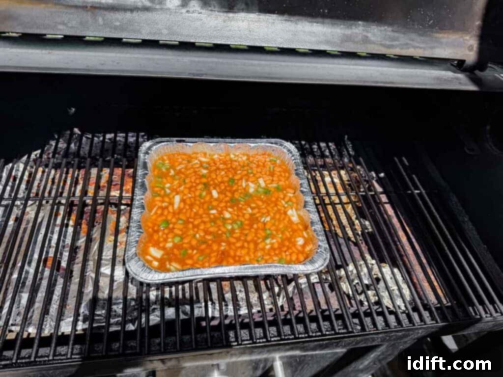 A foil tray filled with baked beans and chopped vegetables sits on a grill inside a barbecue smoker.