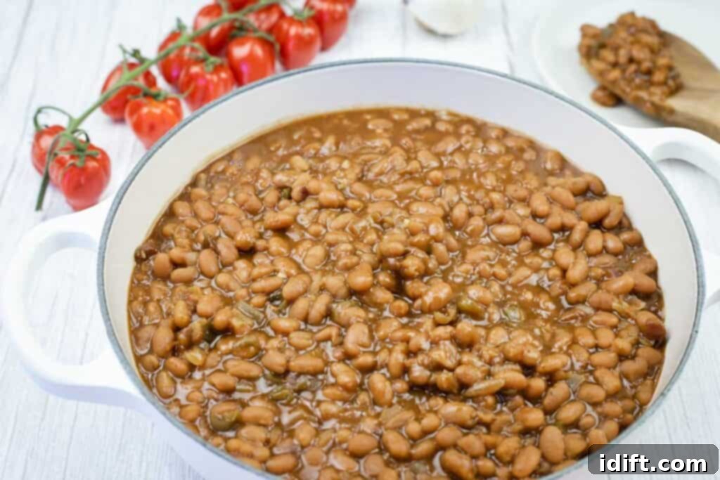 A white pot filled with baked beans in sauce sits on a white surface, with fresh tomatoes and a spoon with beans in the background.