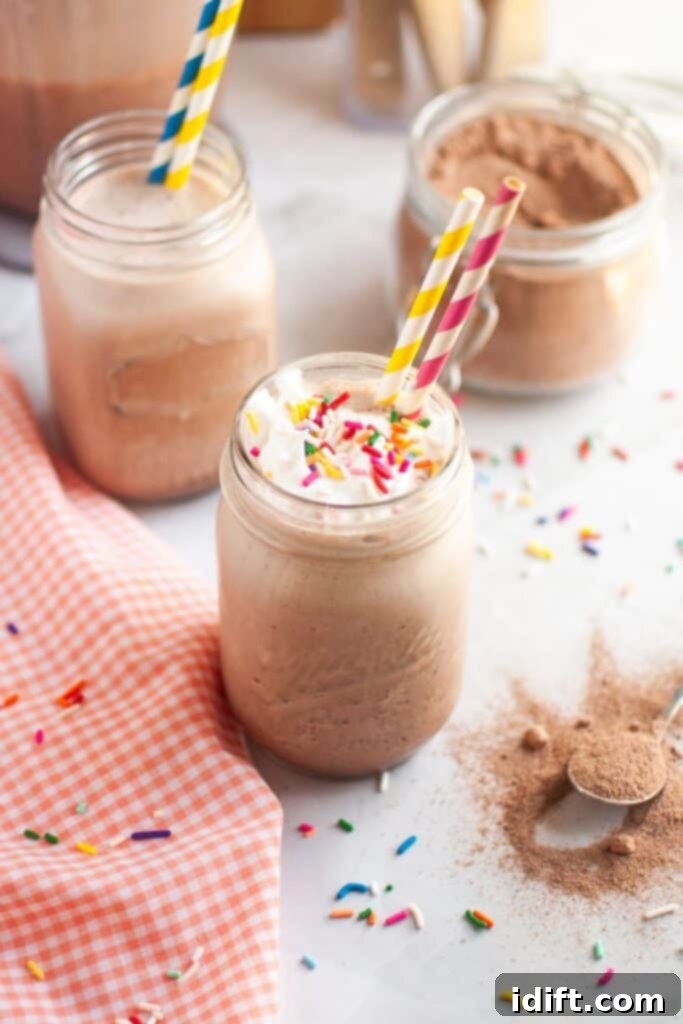 A close-up of a glass of frothy brown frozen hot chocolate, topped with white whipped cream and colorful sprinkles, with a yellow and blue striped straw, on a white countertop with a checkered napkin and another glass in the background. This indulgent dessert drink is perfect for summer.