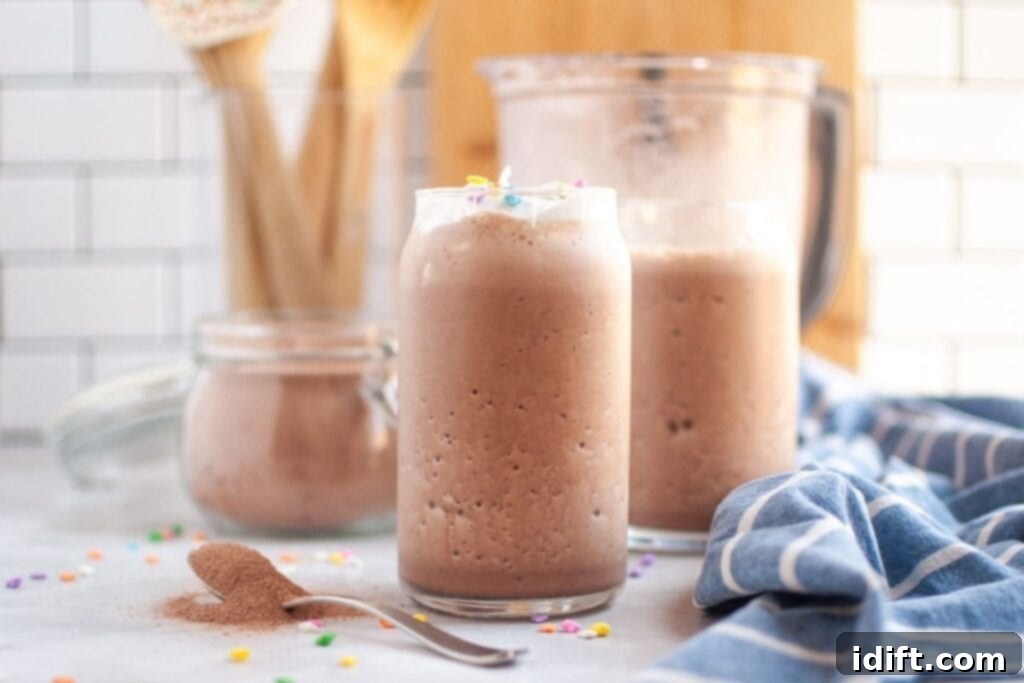 A clear glass filled with frothy brown frozen hot chocolate, topped with a small amount of white whipped cream and colorful sprinkles, on a white countertop with a spoon of cocoa powder, inviting a delicious sip.