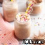 Frosty Chocolate Indulgence 10 A close-up of a glass of frothy brown frozen hot chocolate, topped with white whipped cream and colorful sprinkles, with a yellow and blue striped straw, on a white countertop with a checkered napkin and another glass in the background.