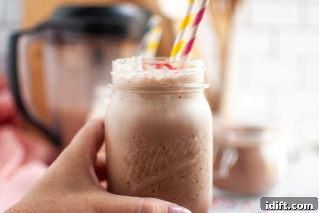 Frosty Chocolate Indulgence 3 A hand with light pink fingernails holds a clear glass of frothy brown frozen hot chocolate topped with white whipped cream and colorful sprinkles.