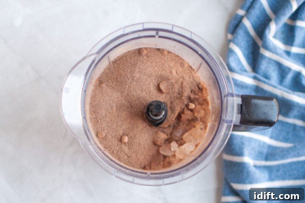 Frosty Chocolate Indulgence 5 Overhead view of a clear food processor bowl containing a brown hot chocolate mix powder and several pieces of crushed ice, on a white surface with a blue and white striped napkin.