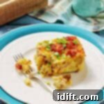 A slice of breakfast casserole topped with bacon and green onions on a white plate with a fork, set on blue and white dishes with a napkin and milk jug in the background.