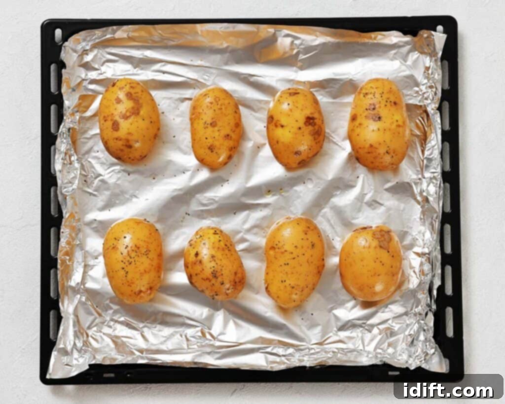 Eight whole, unpeeled potatoes seasoned with pepper are arranged on a foil-lined baking tray, ready for roasting.