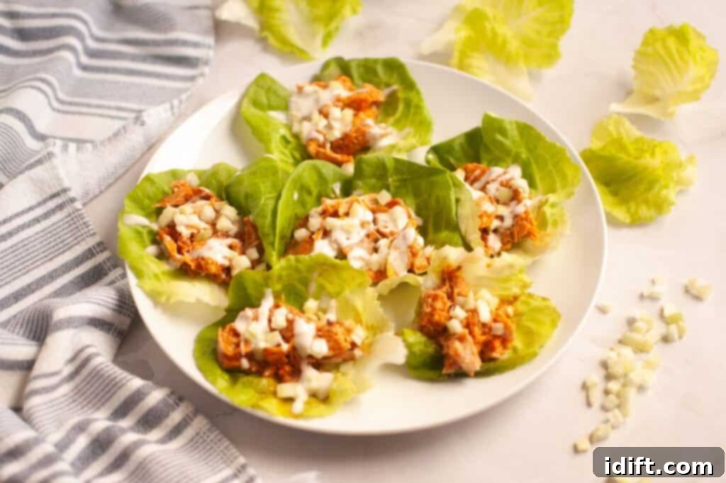 A white plate elegantly displaying six vibrant lettuce cups, each generously filled with shredded buffalo chicken and beautifully topped with crisp diced onions and a luxurious drizzle of creamy sauce. The scene is set on a sophisticated marble surface, accompanied by a stylish striped towel and scattered fresh lettuce leaves.