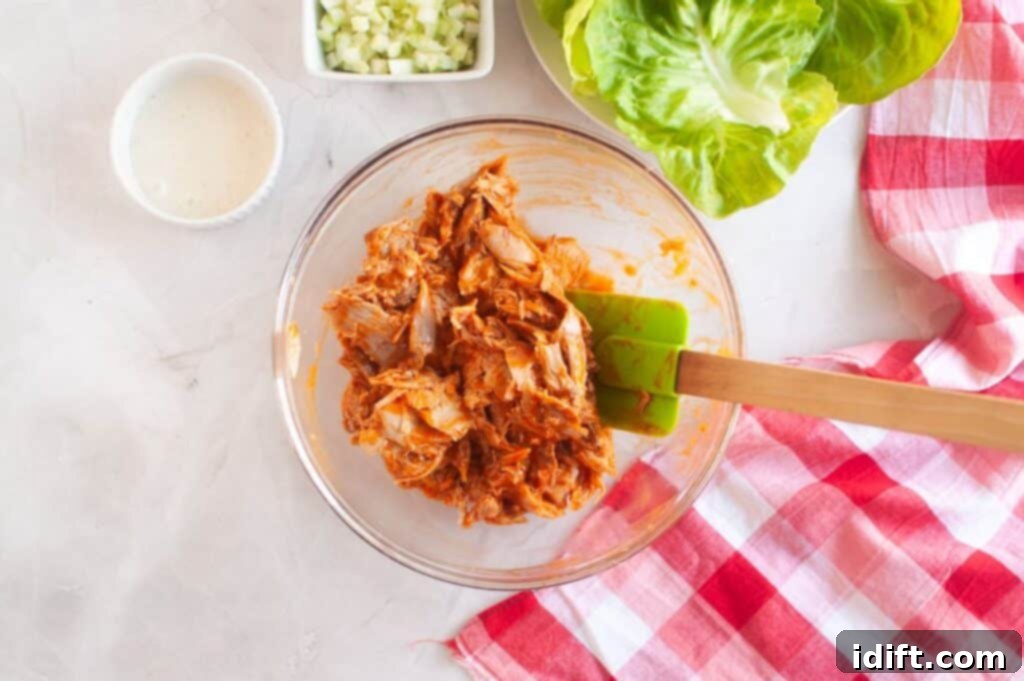 A clear glass bowl filled with shredded chicken mixed generously with buffalo sauce, accompanied by a spatula, fresh lettuce leaves, crisp diced cucumbers, and a bottle of creamy dressing, all set on a clean white surface with a red-and-white checkered cloth.
