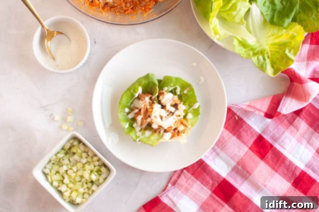 A pristine white plate showcases two beautifully assembled lettuce wraps, generously filled with shredded chicken and elegantly drizzled with creamy white sauce. The setting is completed with a small bowl of fresh lettuce, vibrant diced celery, and a charming red checkered napkin.