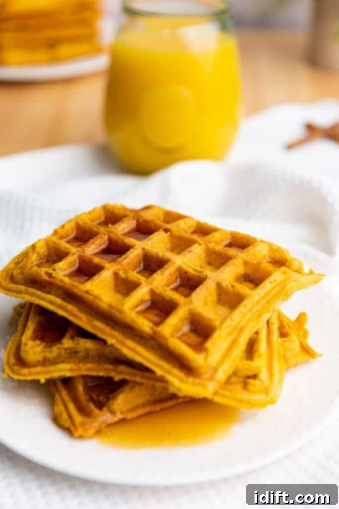 A stack of three square waffles with syrup on a white plate, with a glass of orange juice in the background.