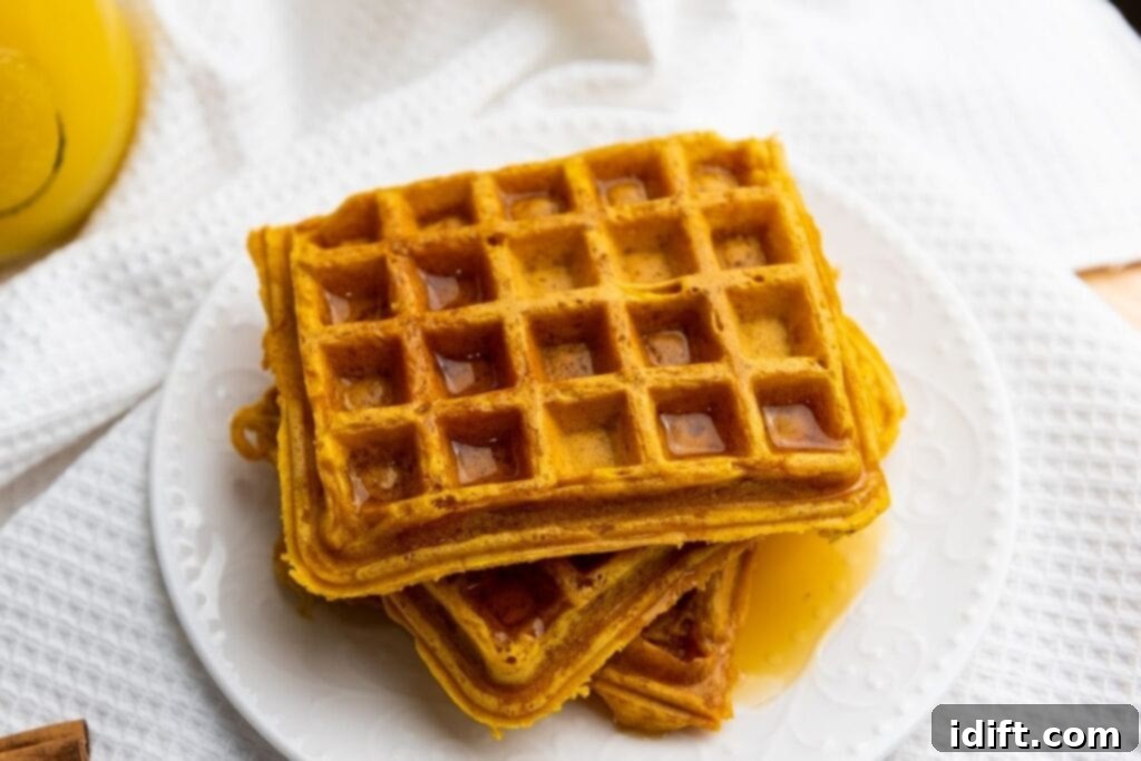 A stack of square waffles with syrup on a white plate, placed on a textured white cloth.