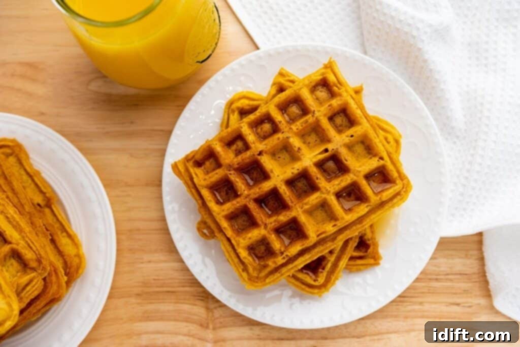 A plate of square waffles with syrup sits on a wooden table next to a glass of orange juice and a white textured napkin.
