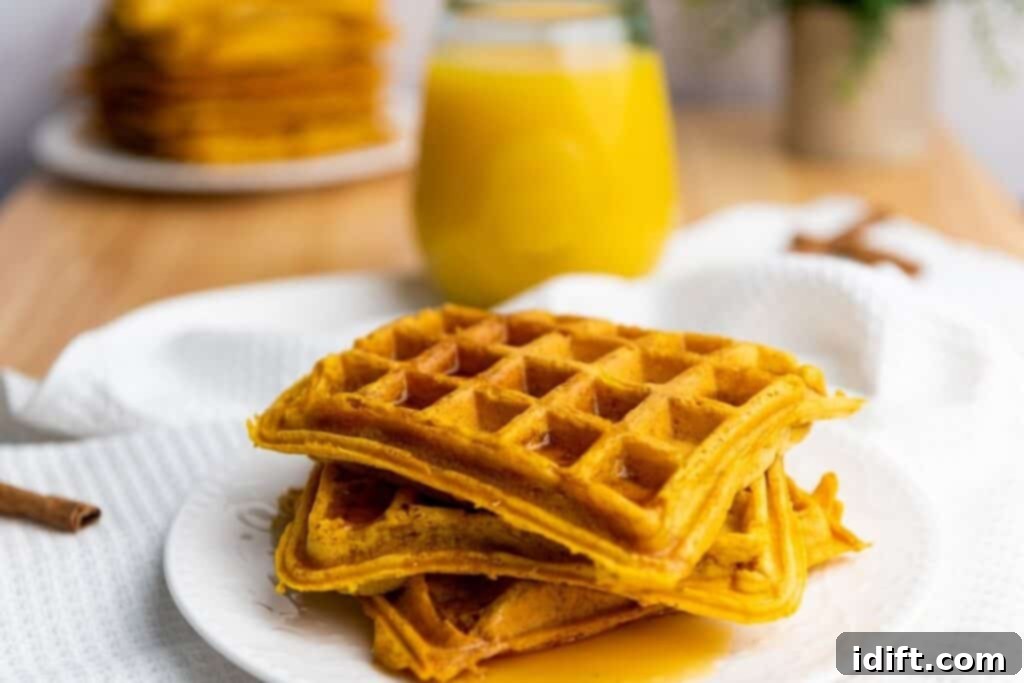 A plate with three stacked waffles drizzled with syrup, with a glass of orange juice and more waffles in the background.