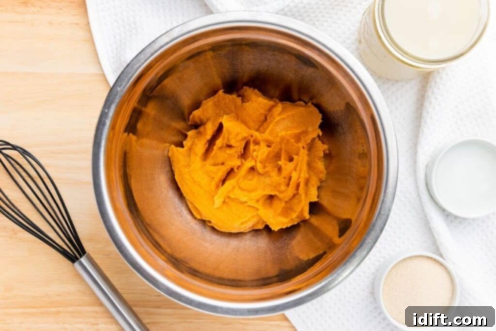 A metal mixing bowl containing pumpkin puree sits on a wooden surface next to a whisk, a jar, a small bowl, and a white towel.