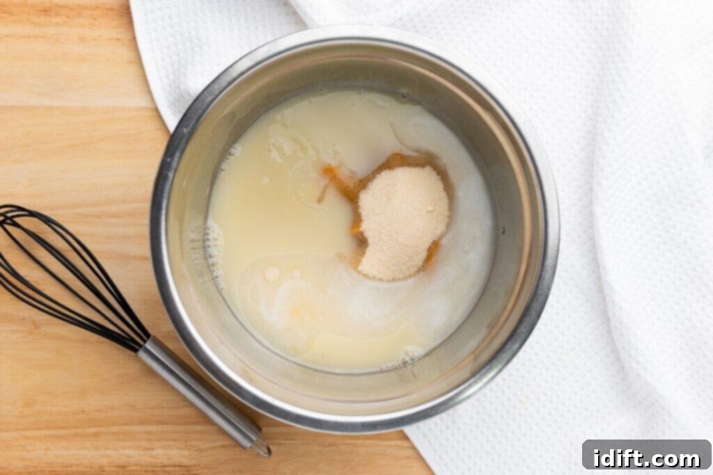 A metal mixing bowl with milk, pumpkin puree, melted butter, and sugar, next to a black wire whisk on a wooden surface with a white towel.