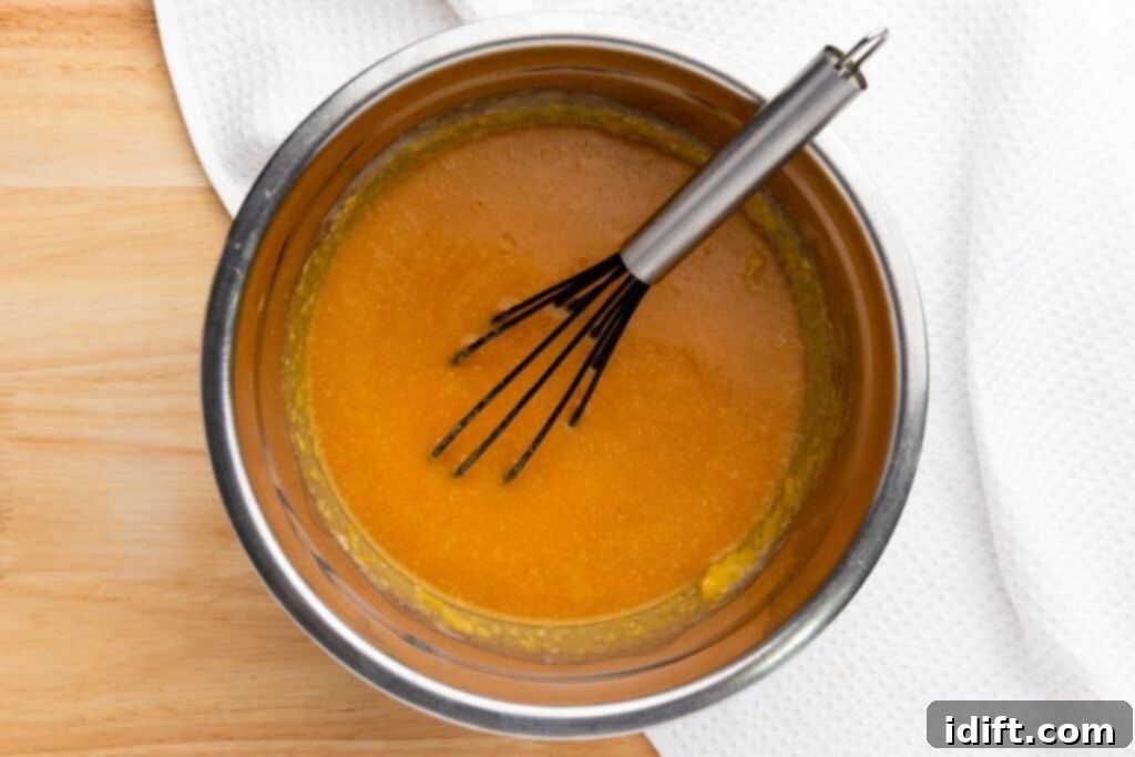 A metal bowl with orange batter being mixed with a whisk, placed on a wooden surface next to a white textured cloth.