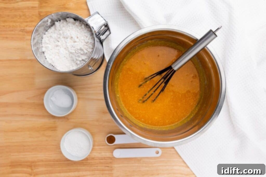 Metal bowl with orange batter and a whisk, flour, small bowls of baking powder and soda, and measuring spoons with cinnamon and salt on a wooden surface next to a white cloth.