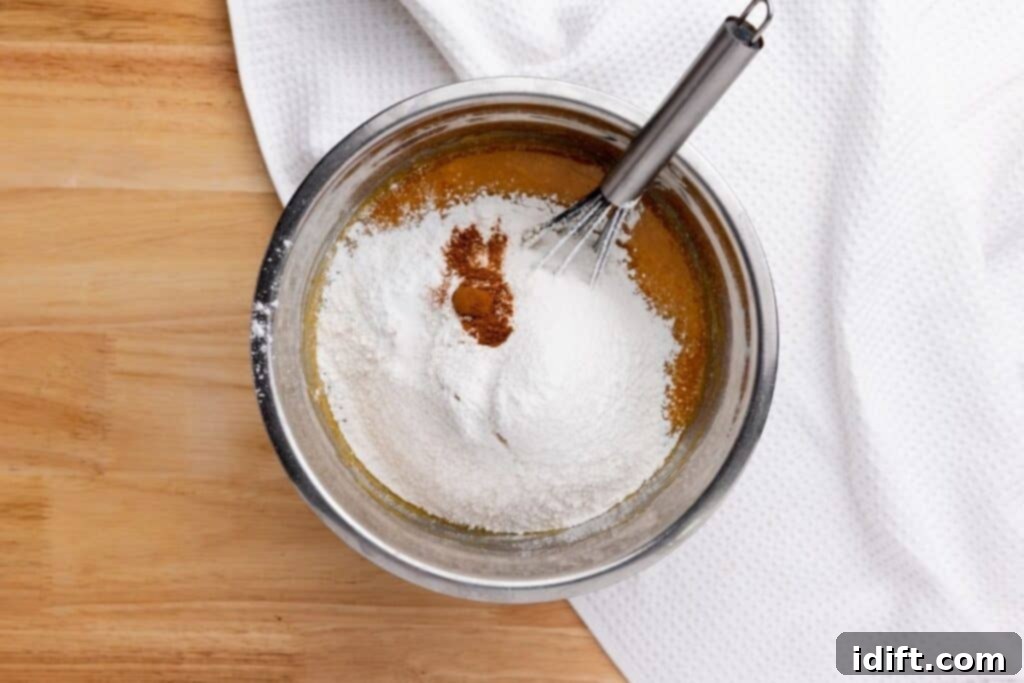 A metal bowl with flour, ground cinnamon, salt, and liquid ingredients being mixed with a whisk on a wooden surface next to a white cloth.
