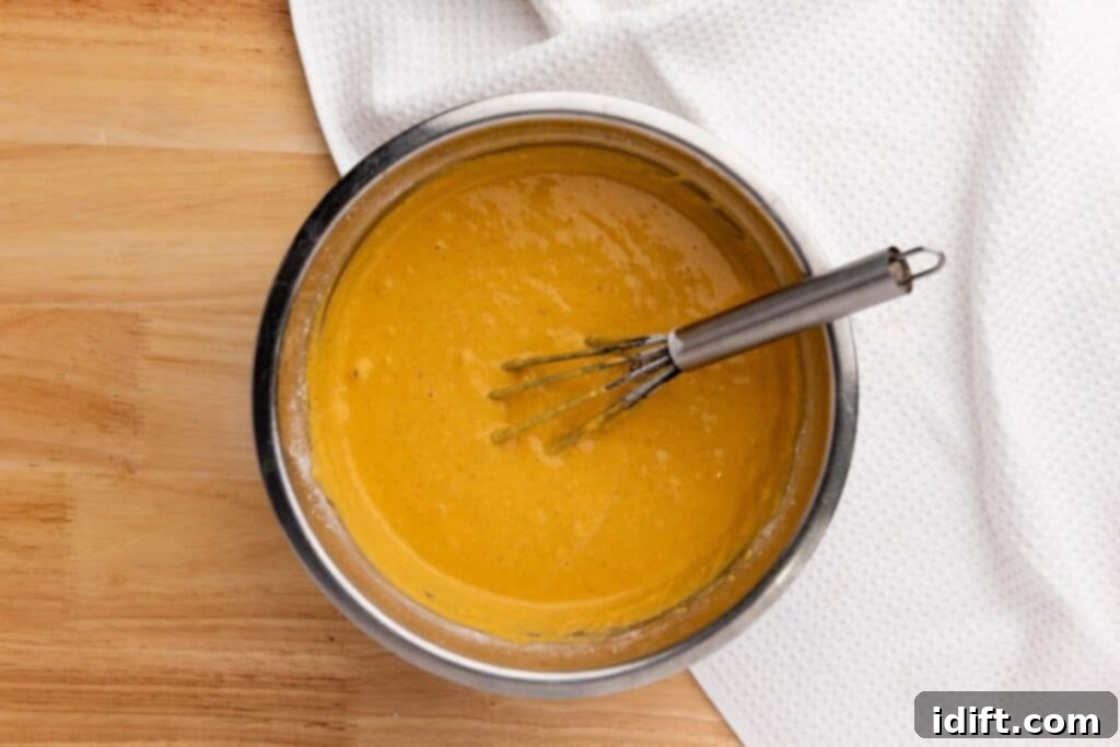 A metal bowl filled with orange batter and a whisk sits on a wooden surface next to a white textured cloth.