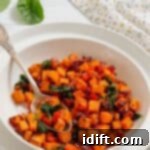 Golden Sweet Potato and Spinach Hash 6 A white bowl filled with a sweet potato hash containing diced sweet potatoes, red onions, spinach, and red bell peppers, with a spoon resting inside.