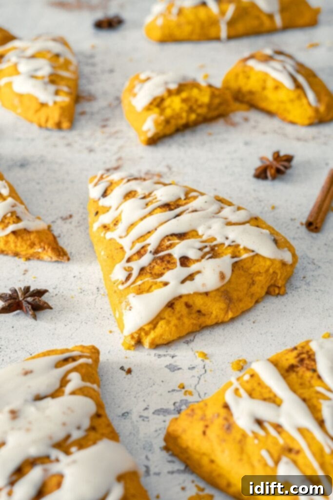 Pumpkin scones with white icing are arranged on a light surface, with some whole and one halved, surrounded by cinnamon sticks and star anise.
