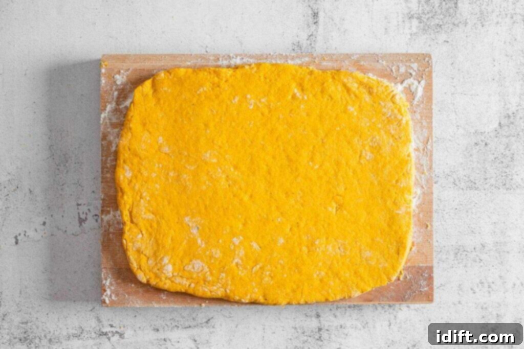 A rectangular sheet of orange dough is rolled out on a floured wooden board, placed on a light-colored textured surface.