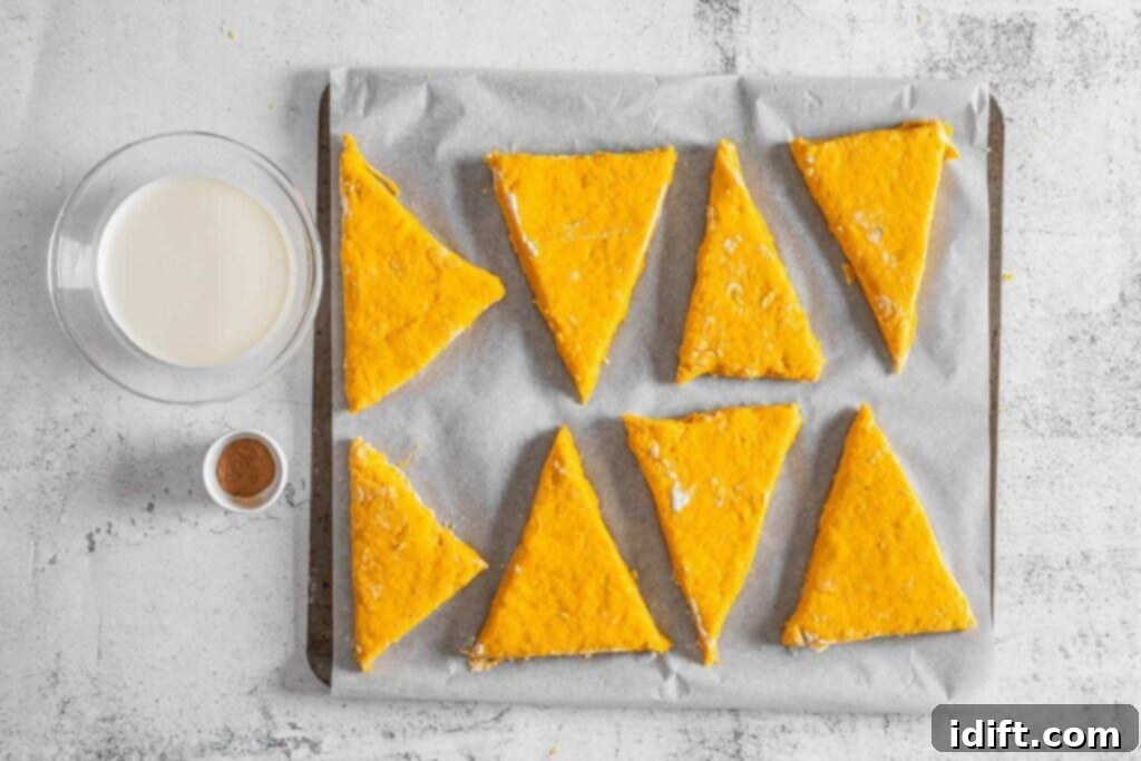 Eight unbaked triangular scones on a parchment-lined baking tray, with a small bowl of pumpkin pie spice and a glass cream on the side.