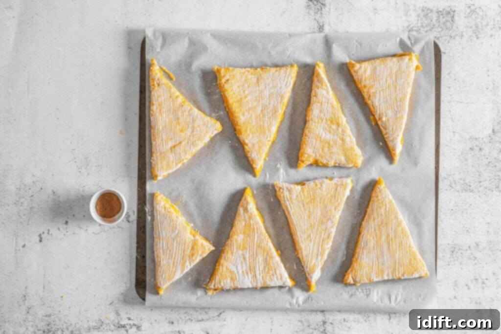 Eight unbaked, triangular scones with cream on parchment paper, placed on a baking sheet; a small container of pumpkin pie spice sits beside them.