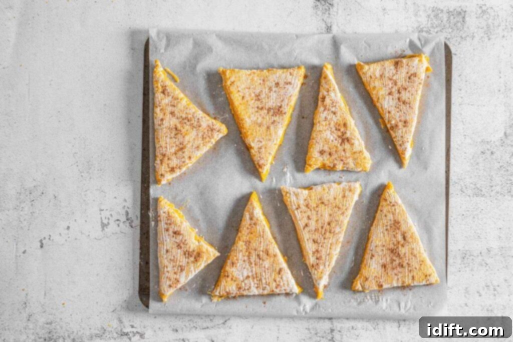 Eight triangular pieces of uncooked pastry, dusted with pumpkin pie spice, arranged on a sheet of parchment paper atop a baking tray.