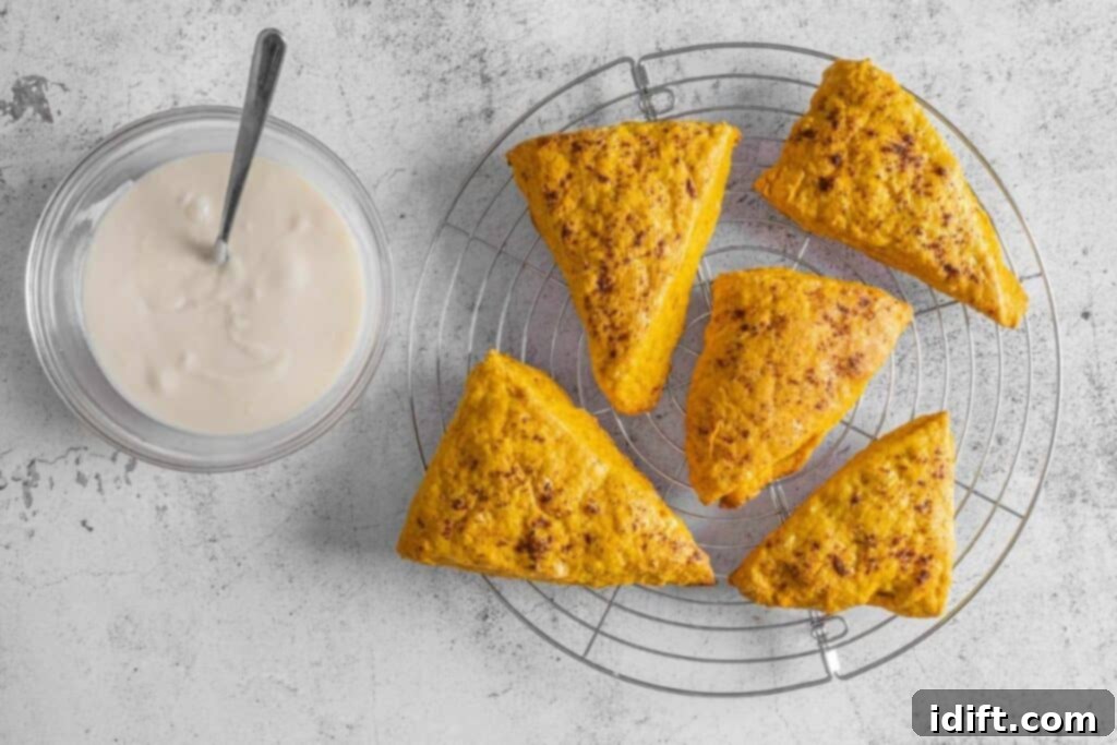 Five triangular scones rest on a round wire cooling rack next to a glass bowl of glaze with a spoon on a light gray surface.