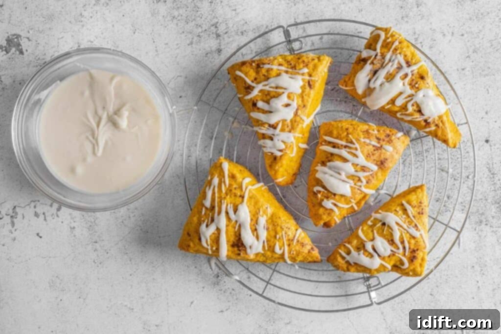 Five triangular scones with white glaze are on a round cooling rack next to a glass bowl filled with glaze, all on a light-colored surface.