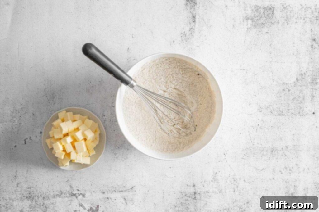 A bowl of dry ingredients mix with a whisk inside next to a small bowl of cubed butter on a light textured surface.