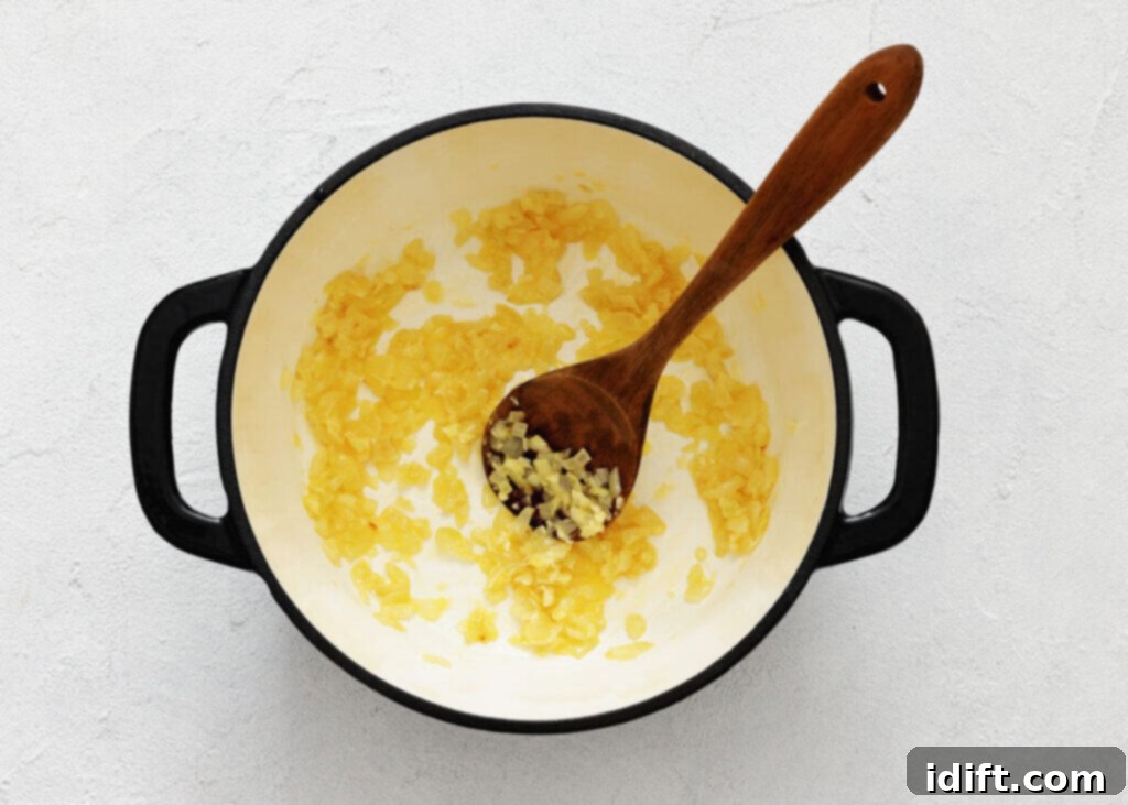 Finely chopped onions are being gently sautéed in a white cooking pot, stirred with a wooden spoon. The onions are becoming translucent, indicating they are softening and releasing their natural sweetness, forming the aromatic foundation for the soup.