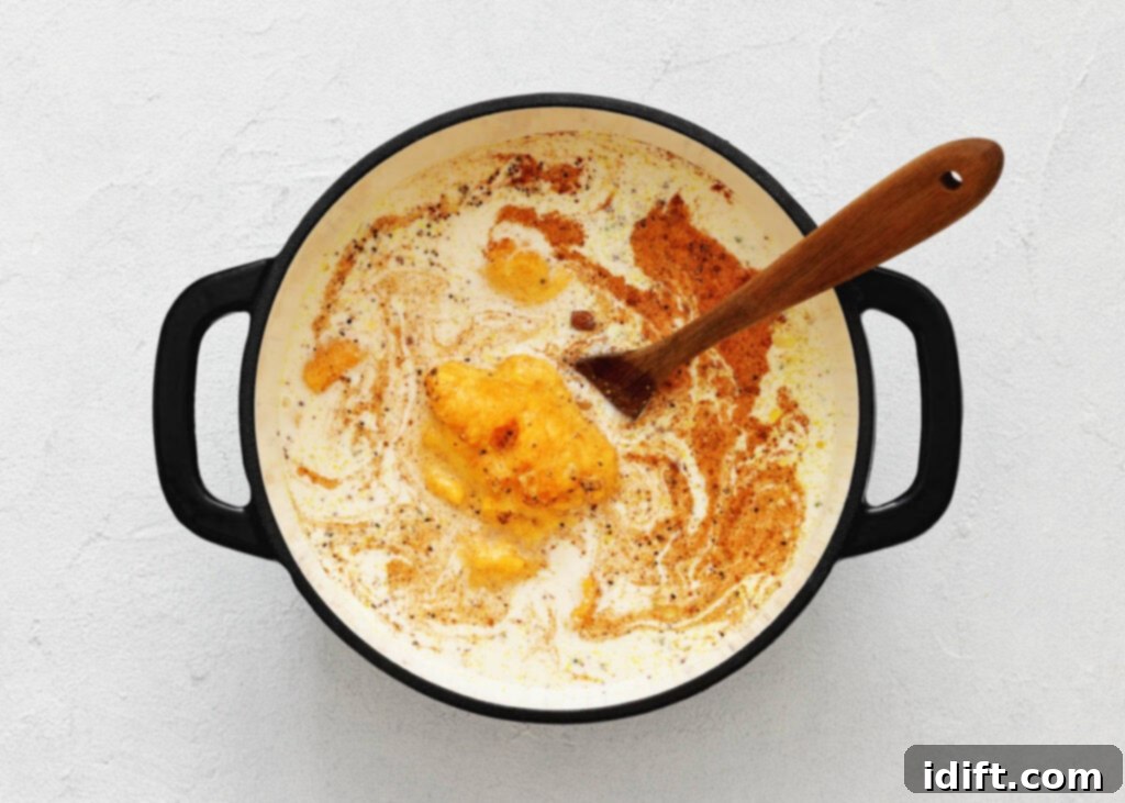 A black cooking pot on a white surface contains a creamy mixture of liquid, melted butter, and various spices, being stirred with a wooden spoon. The ingredients are in the process of being blended to create a smooth and cohesive base for the soup.