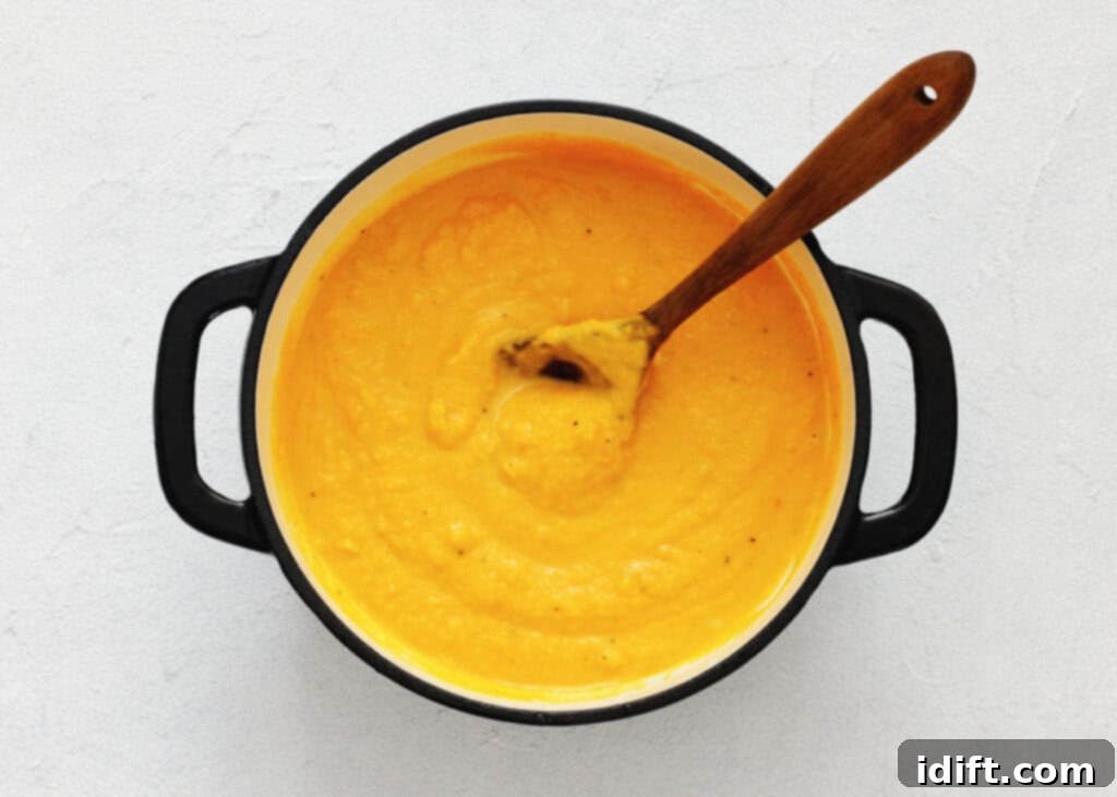 A pot of rich, thick, and creamy orange soup is shown from a top-down perspective, resting on a light-colored surface. A wooden spoon is placed inside the pot, suggesting the soup has just been prepared or is ready for serving, highlighting its smooth texture.