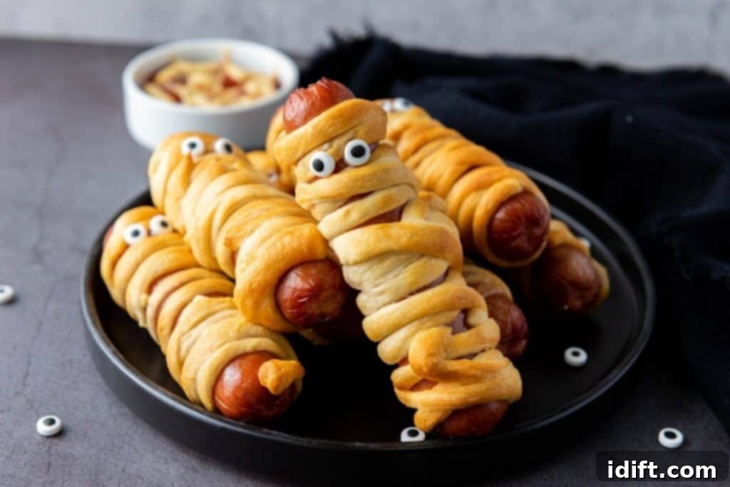 A plate featuring several perfectly air-fried mummy hot dogs, each wrapped in a golden-brown dough and adorned with candy eyes, creating a charmingly spooky effect. A small bowl of dipping sauce, blurred in the background, completes the festive presentation.