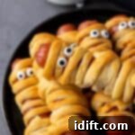 Several hot dogs wrapped in crescent dough to look like mummies, with candy eyes added, are arranged on a black plate. A small bowl of dipping sauce is in the background.