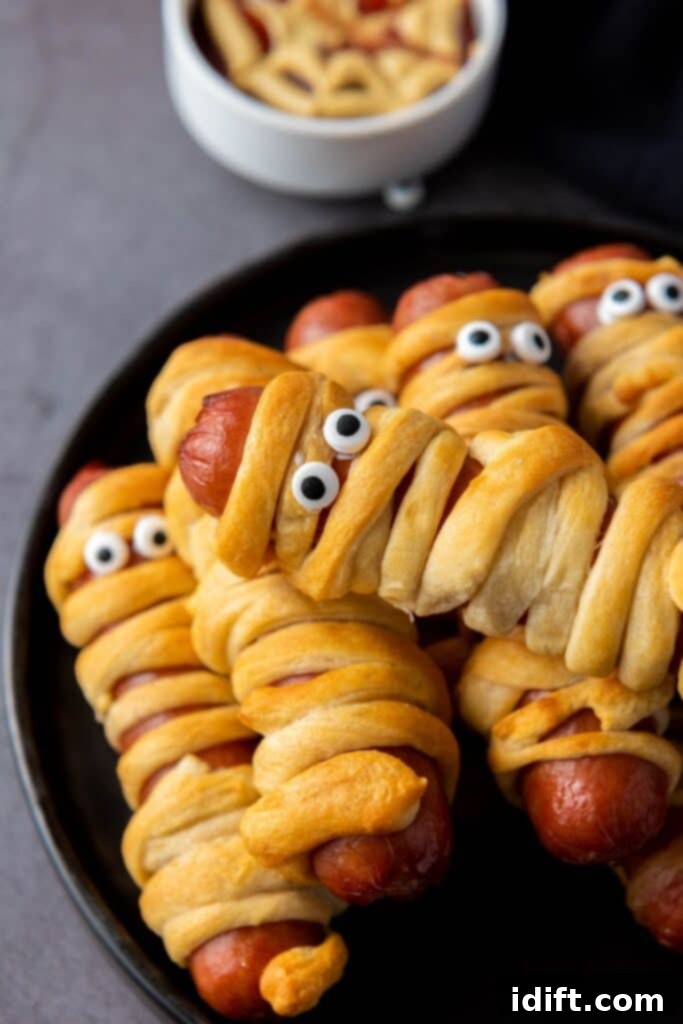 Several air-fried hot dogs, artfully wrapped in crescent dough to resemble mummies, are arranged on a stylish black plate. Each mummy dog features two edible candy eyes, adding to their playful Halloween theme. A small, out-of-focus bowl of dipping sauce is visible in the background.
