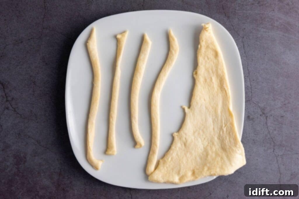 A white plate displays strips of uncooked crescent dough alongside an uncut triangle of dough, ready for wrapping. The arrangement is on a dark countertop, highlighting the dough's texture.